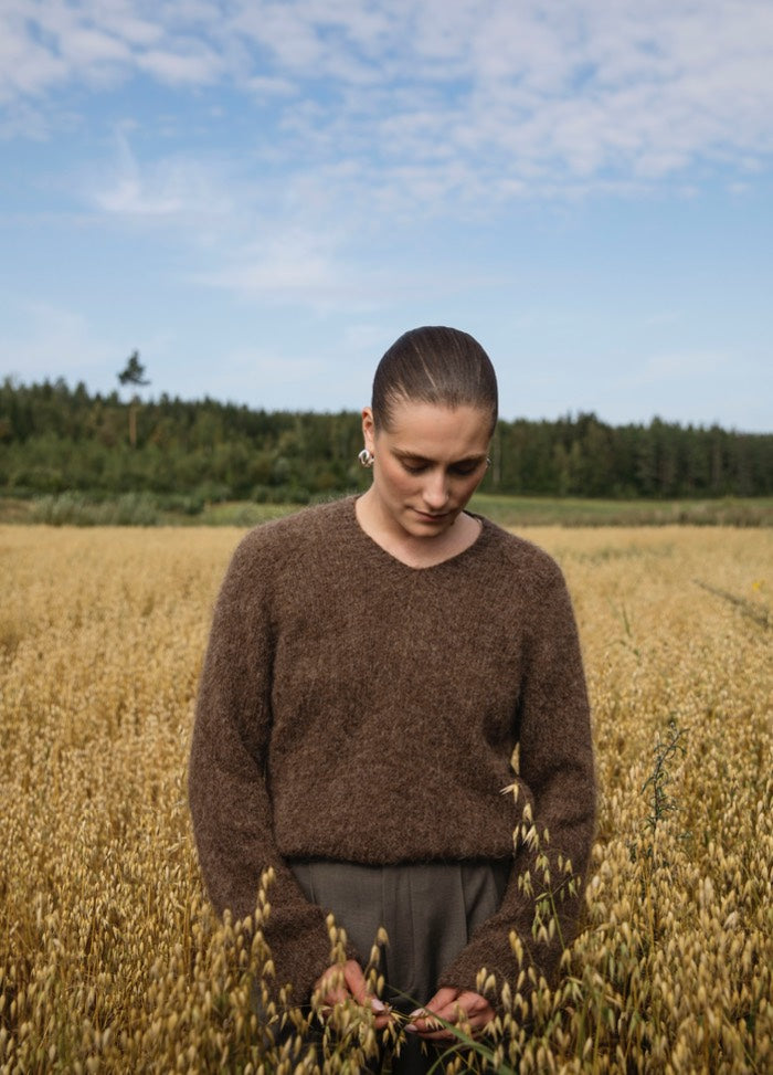 Sally Sweater': Brown sweater worn in a field of golden crops with forest and blue sky in the background.