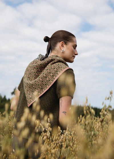 Walled Garden Scarf': Floral scarf with pink border, draped over shoulders in a field, worn with green top and hair in a bun.