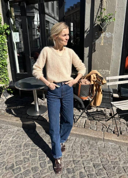 A person in a beige sweater and blue jeans stands on a cobblestone street, with hands in pockets. Sunlight casts sharp shadows. A café and plants are nearby.