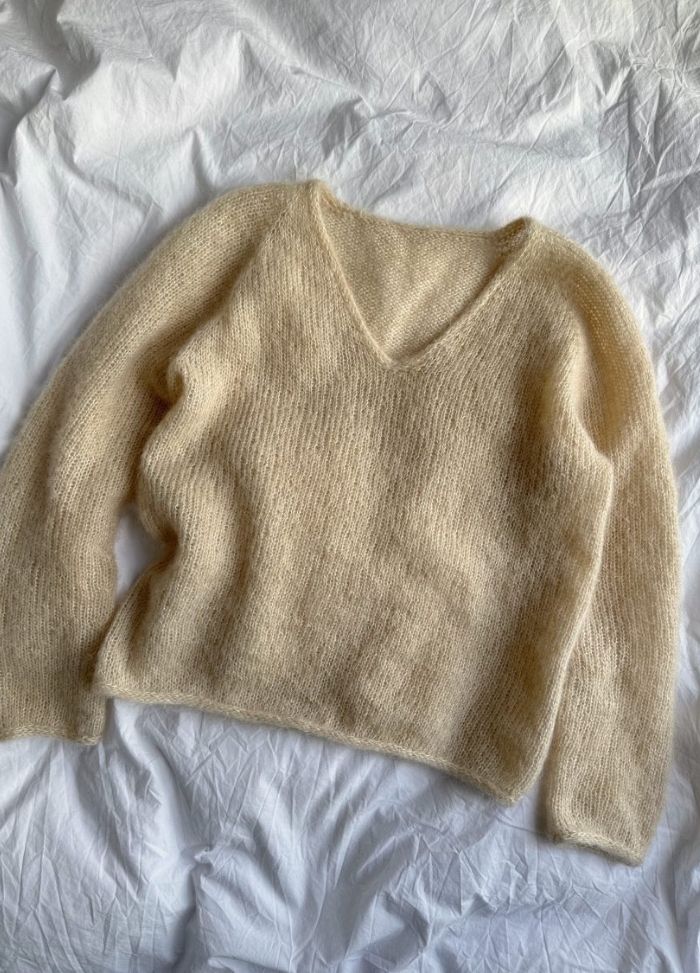 A soft, cream-coloured fuzzy sweater with a V-neck and long sleeves, lying flat on a textured white surface, conveying warmth and coziness.