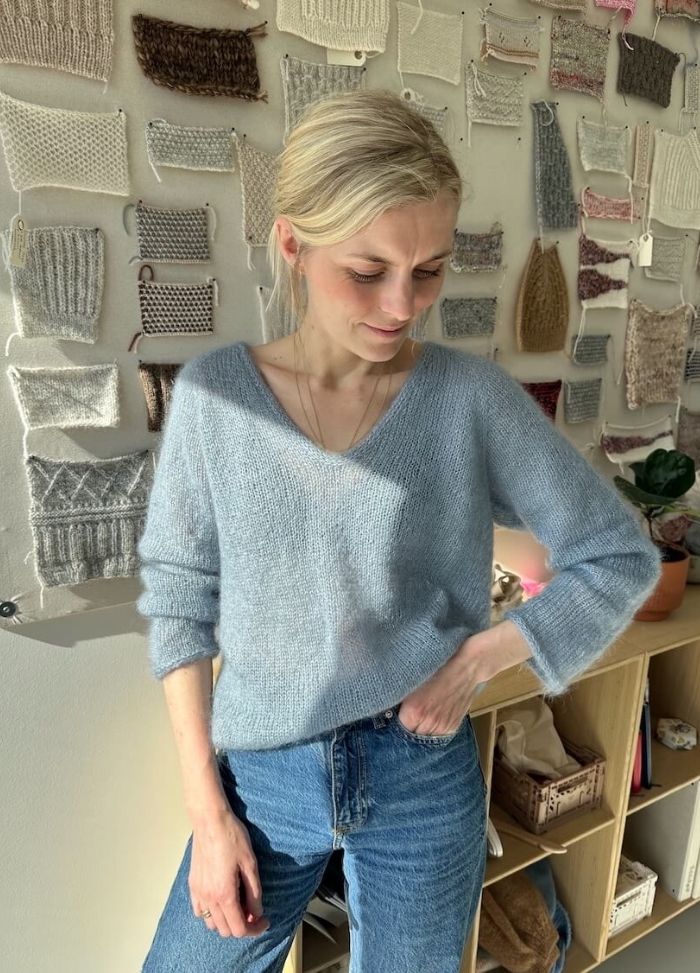 A person in a light blue sweater and jeans stands in a sunlit room, smiling slightly. Behind them, a wall displays various knitted swatches. The mood is calm and creative.