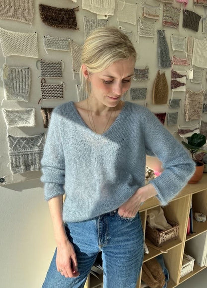 A person in a light blue sweater and jeans stands in a sunlit room, smiling slightly. Behind them, a wall displays various knitted swatches. The mood is calm and creative.
