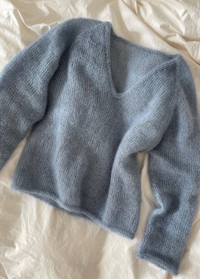 A soft, blue knit sweater with long sleeves and a V-neck lies on a rumpled, off-white fabric background, conveying a cozy and warm feel.