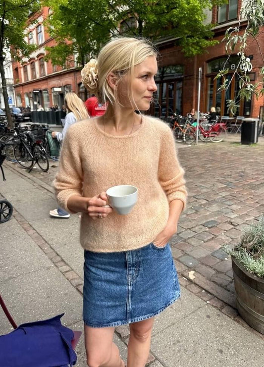 A woman stands on a cobblestone street holding a white mug, wearing a peach sweater and denim skirt. She gazes away, with bicycles and trees nearby.