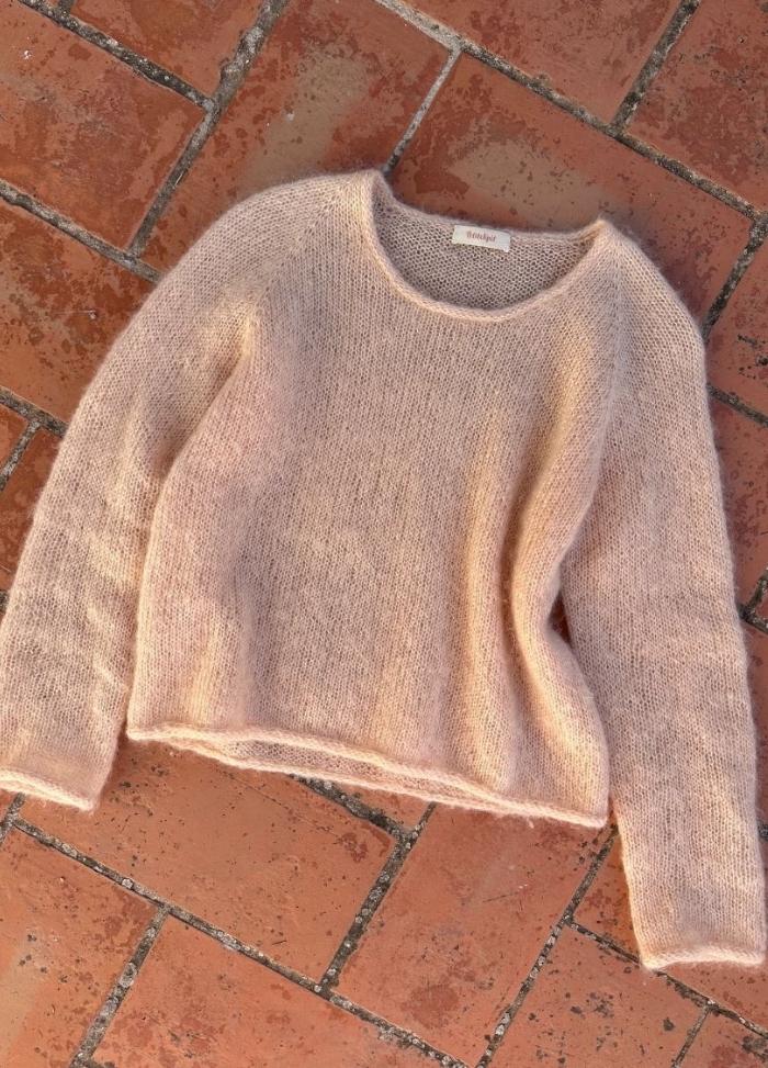 A soft pink, fuzzy sweater lies on a terracotta brick floor. The warm sunlight enhances its cozy texture, creating a relaxed and inviting feel.