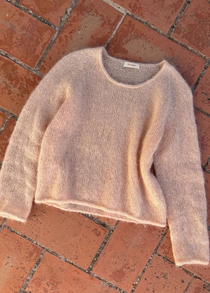 A soft pink, fuzzy sweater lies on a terracotta brick floor. The warm sunlight enhances its cozy texture, creating a relaxed and inviting feel.
