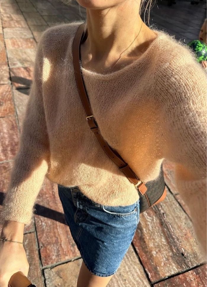 Person in a fuzzy, beige sweater and denim skirt on a sunlit stone pavement. Brown crossbody bag, casual, warm tone, outdoor setting.