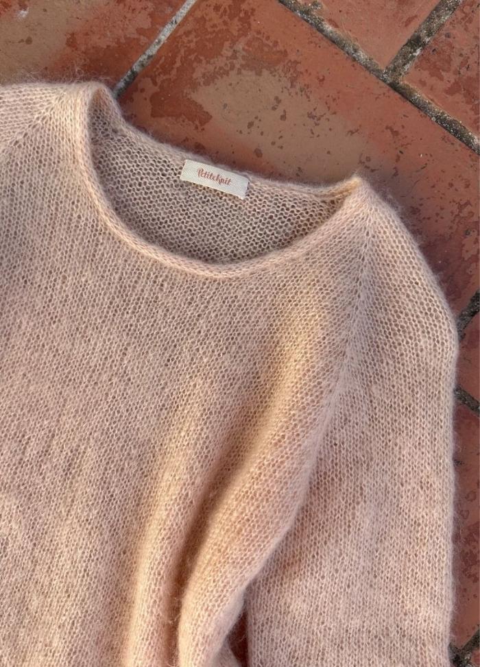 A soft, beige mohair sweater with a round neck lies on terracotta tiles. Its fluffy texture and pastel hue create a cozy, warm ambiance.
