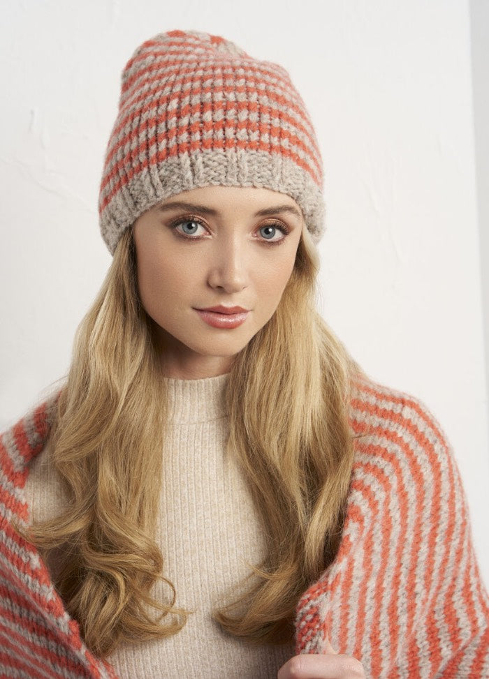A woman with long blonde hair wears a red and gray knit beanie and matching striped shawl. She gazes softly at the camera, conveying warmth and coziness.