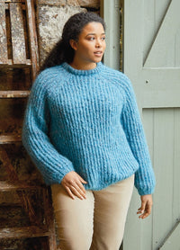 Woman in a cozy blue knit sweater and beige pants stands against a rustic backdrop of a wooden ladder and door, exuding a calm and thoughtful vibe.