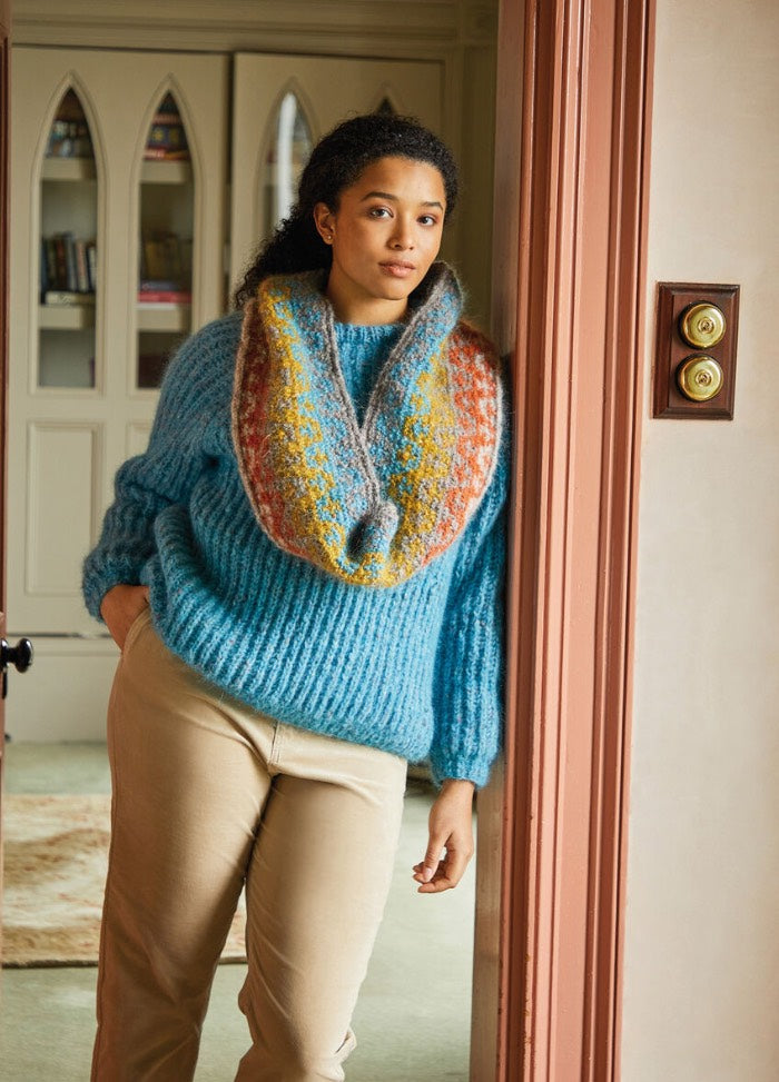 A person in a cozy blue sweater and colorful scarf leans casually against a door frame, exuding warmth and comfort. Decorative paneling is visible behind them.