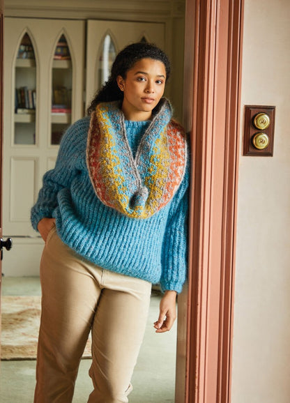 A person in a cozy blue sweater and colorful scarf leans casually against a door frame, exuding warmth and comfort. Decorative paneling is visible behind them.