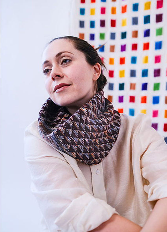 Model wearing the Cygnus Cowl, a textured knit in interwoven brown and beige tones, styled over a light shirt with a colorful grid backdrop.