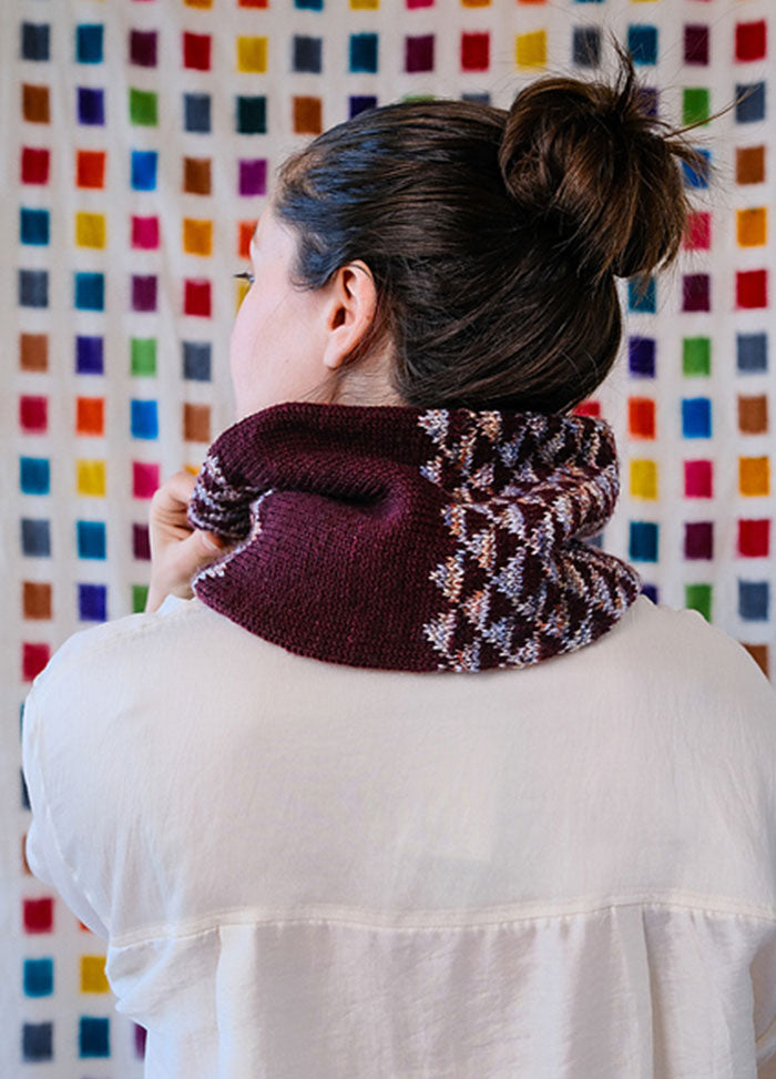 Model wearing the Cygnus Cowl, a maroon knit with an intricate white and multicolor pattern, set against a colorful grid backdrop.
