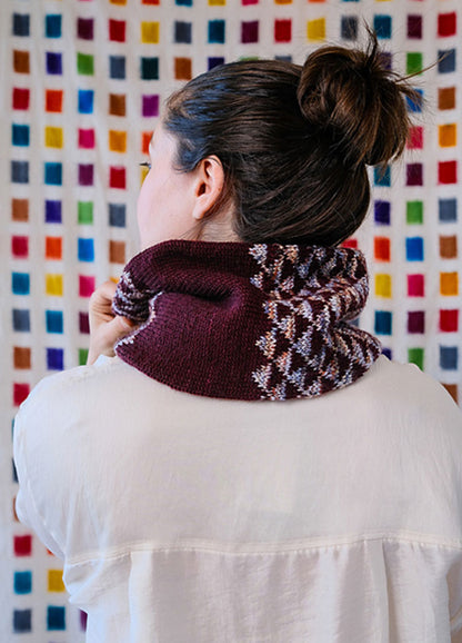 Model wearing the Cygnus Cowl, a maroon knit with an intricate white and multicolor pattern, set against a colorful grid backdrop.