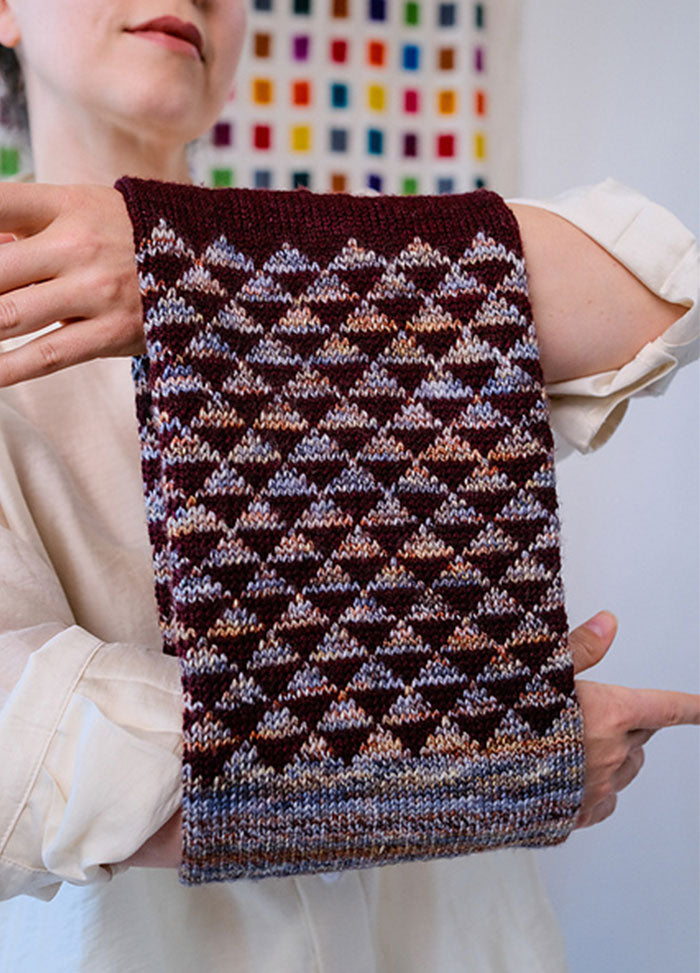 Model wearing the Cygnus Cowl, a maroon knit with geometric diamond patterns in lighter shades, set against a colorful grid backdrop.