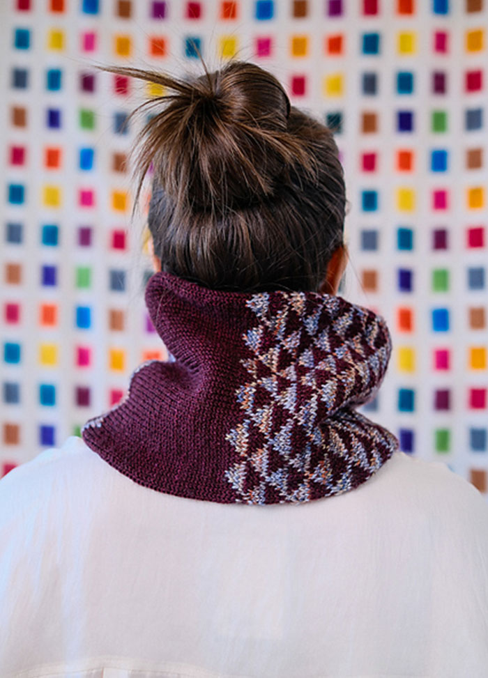 Model wearing the Cygnus Cowl, a deep burgundy knit with interlocking diamond motifs in contrasting colors, set against a colorful grid backdrop.