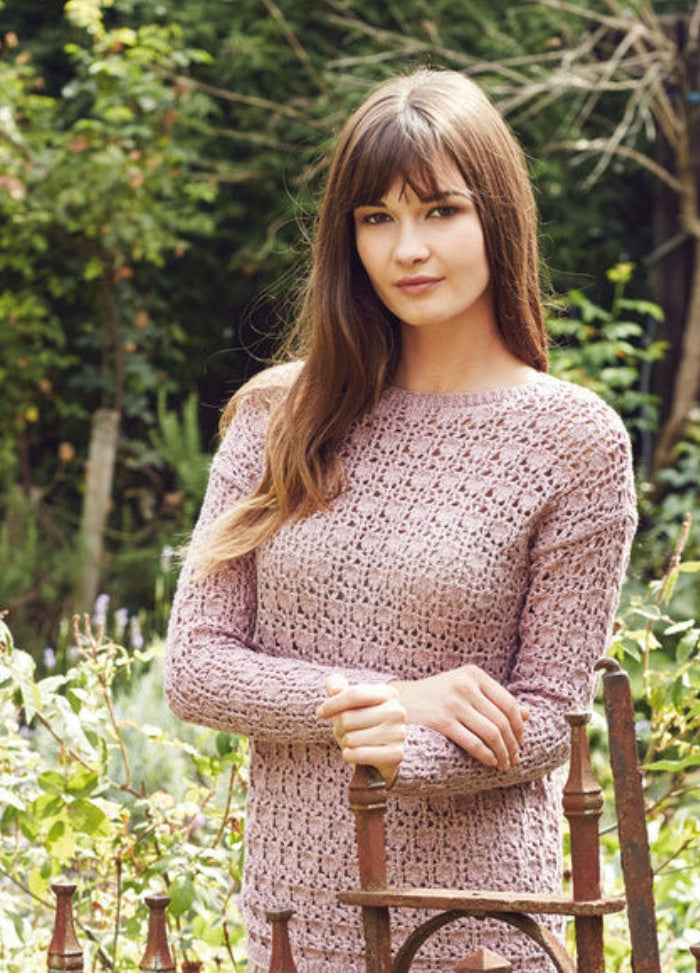 Model wearing the Dawa sweater from the Rowan Softyak DK Collection by Marie Wallin, a light pink long-sleeved pullover with an open-knit textured pattern, photographed in a garden setting with greenery and iron railing.