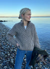 A woman in a beige knit sweater and jeans stands on a rocky beach, holding a jacket. The calm sea and clear blue sky create a serene and peaceful atmosphere.