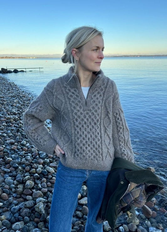 A woman in a beige knit sweater and jeans stands on a rocky beach, holding a jacket. The calm sea and clear blue sky create a serene and peaceful atmosphere.