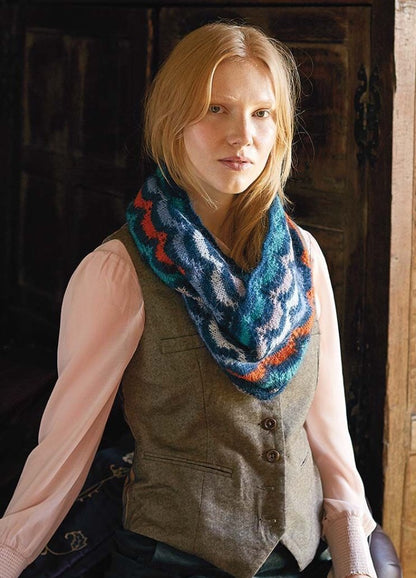 A person with long hair wears a colorful, zigzag-patterned knitted scarf, a pink blouse, and a gray vest. They stand indoors, exuding a calm demeanor.