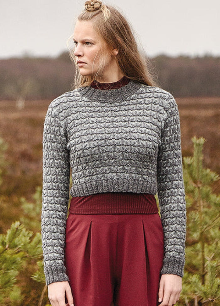 Person wearing the Dales Sweater, a textured grey knit with long sleeves, paired with maroon pants in an open field setting.