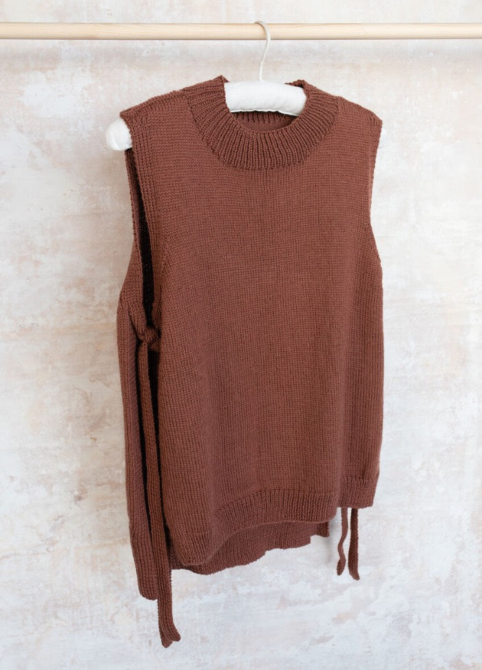 Brown sleeveless knitted vest with side ties hangs on a wooden hanger against a textured, light beige wall. The tone is warm and cozy.