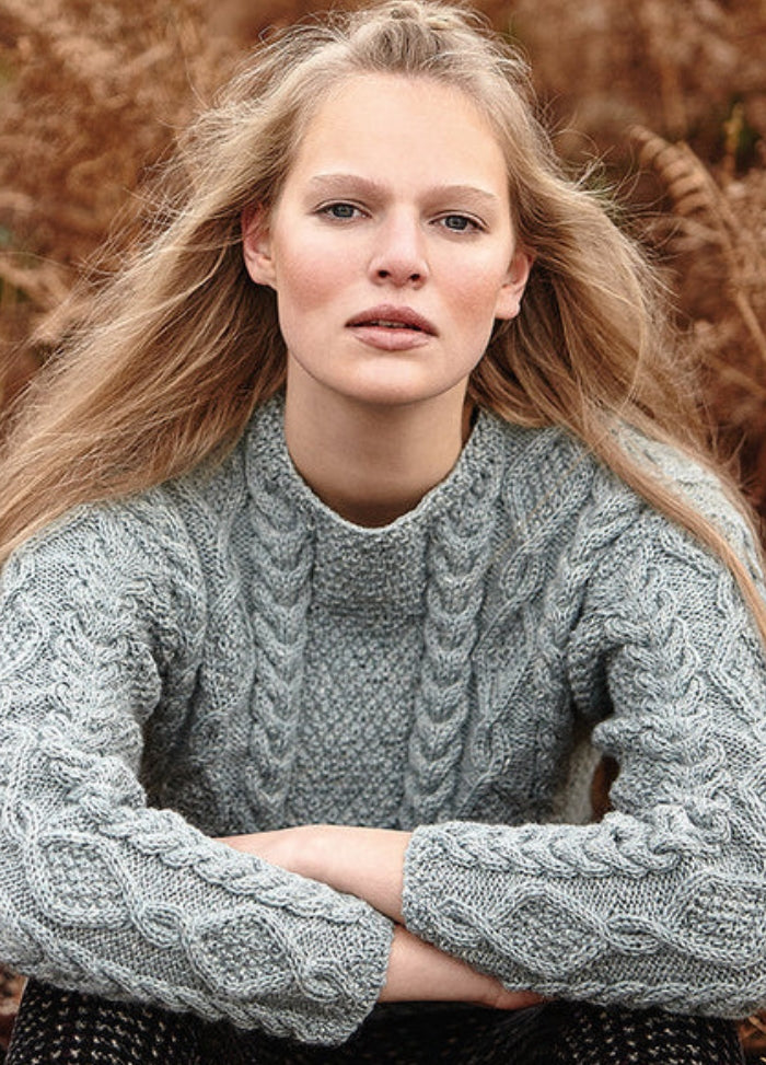 Person wearing the Dartmoor Sweater, a grey cable-knit design with detailed patterns on sleeves and front, photographed outdoors with brown foliage.