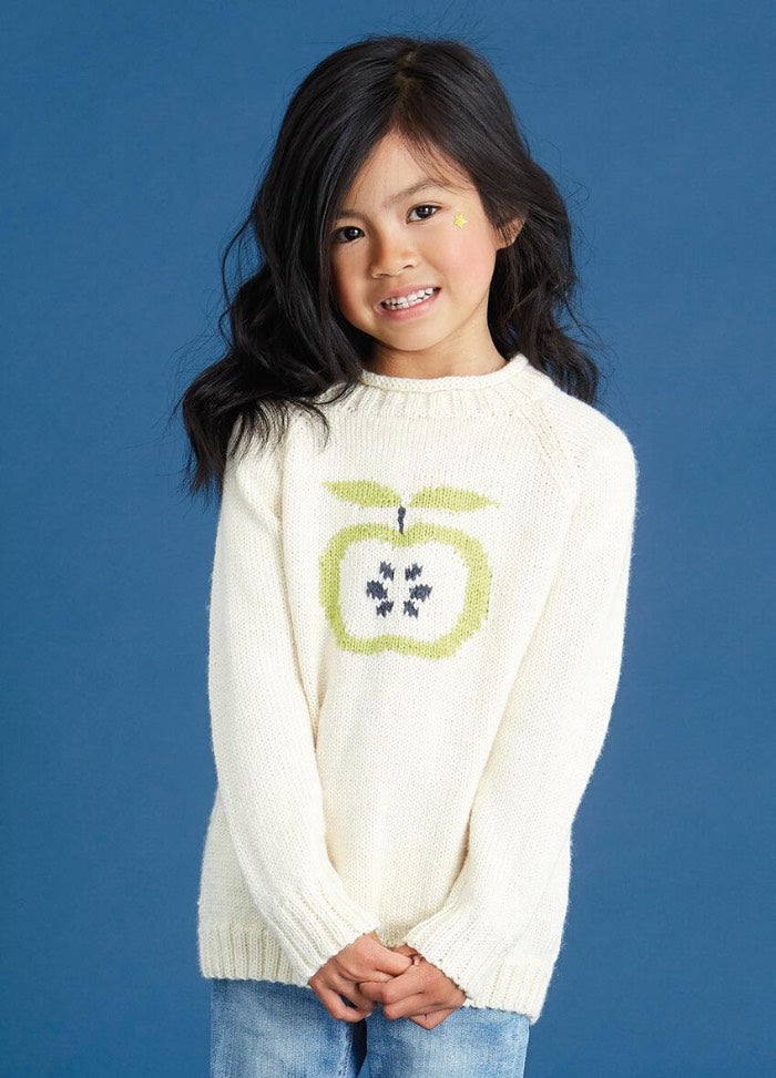 Young girl with long black hair smiles softly, wearing a cream sweater featuring a green apple design, and light blue jeans, against a blue background.