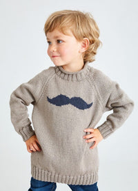 Young child with blond hair wearing a light brown sweater featuring a large navy mustache design, hands on hips, smiling confidently. Light background.