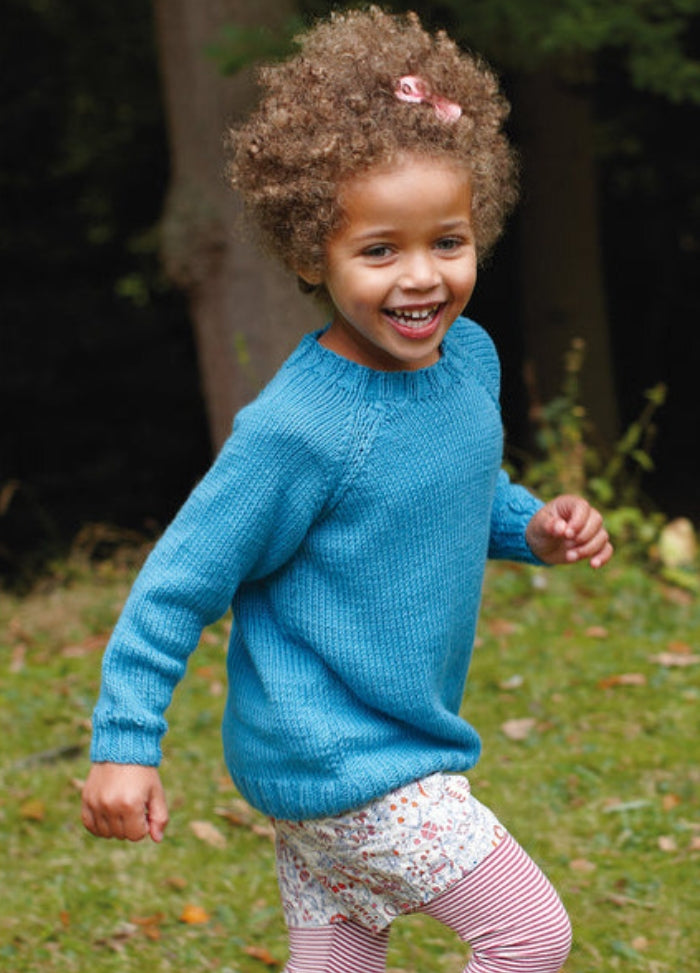 Child in blue Deacon Sweater with patterned shorts and striped leggings, outdoors.