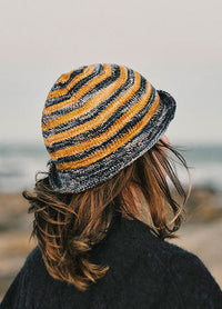 Back view of a striped knit hat, the Delmira Hat, in yellow, black, and gray, worn outdoors near a rocky shoreline.