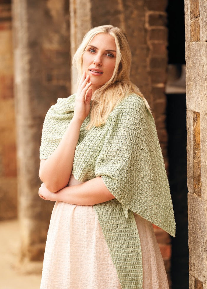 A woman with blonde hair stands against a rustic stone wall, wearing a light green shawl and beige dress, exuding a calm, serene expression.
