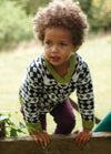 Child in Diamond Sky Sweater with black and white diamonds and green trim, leaning on wood.