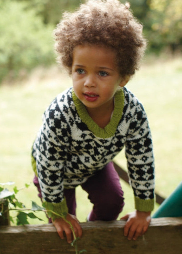 Child in Diamond Sky Sweater with black and white diamonds and green trim, leaning on wood.