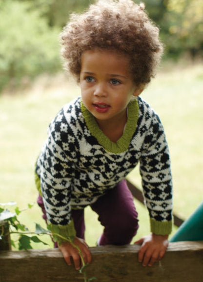 Child in Diamond Sky Sweater with black and white diamonds and green trim, leaning on wood.