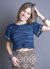A smiling woman in a blue knitted crop top with beaded sleeves and a patterned skirt. She is touching her hair, wearing bracelets, and exudes a joyful, relaxed vibe.
