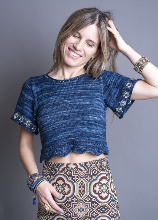 A smiling woman in a blue knitted crop top with beaded sleeves and a patterned skirt. She is touching her hair, wearing bracelets, and exudes a joyful, relaxed vibe.
