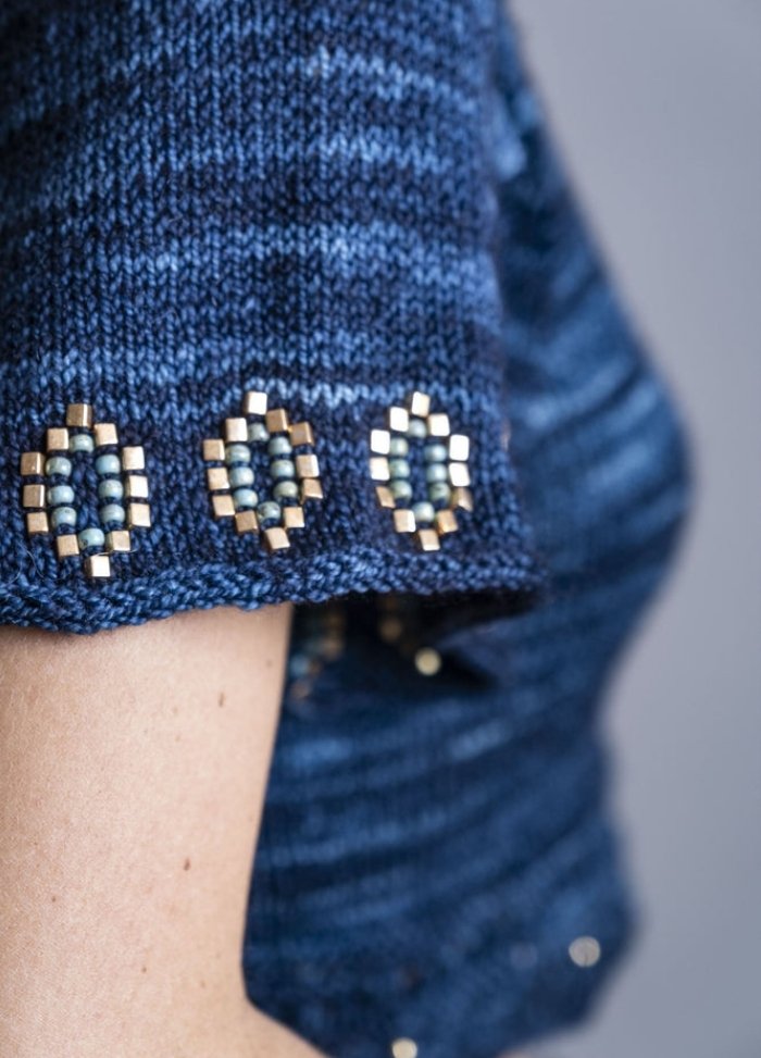 Close-up of a blue knitted sweater sleeve adorned with intricate beading. Gold and blue beads form a geometric pattern, creating a handcrafted, elegant look.