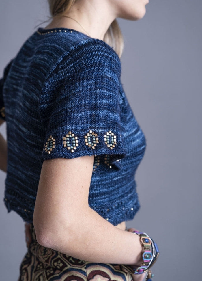 Woman in a blue knit top with detailed geometric trim on sleeves and hem, wearing a patterned skirt and bracelets, against a gray background.