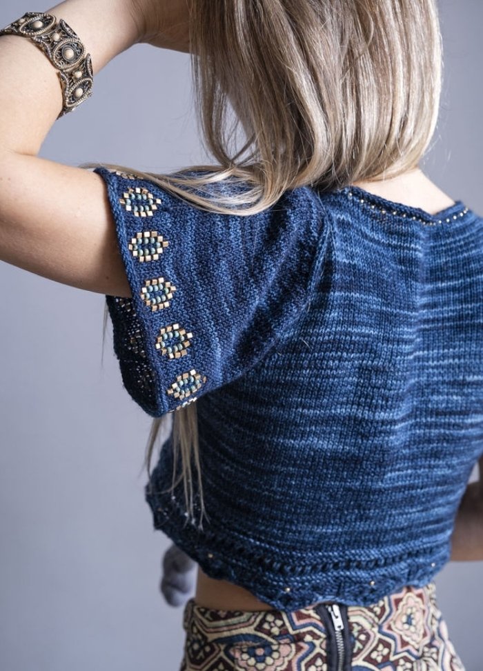 A woman with long blonde hair is seen from behind, wearing a navy knit top with intricate beadwork on the sleeves. She has a decorative bracelet and a patterned skirt, creating a bohemian style.