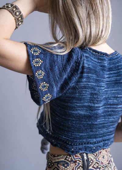 A woman with long blonde hair is seen from behind, wearing a navy knit top with intricate beadwork on the sleeves. She has a decorative bracelet and a patterned skirt, creating a bohemian style.