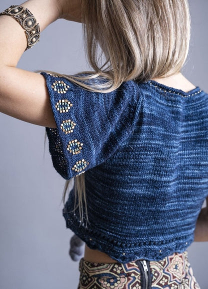 A woman with long blonde hair is seen from behind, wearing a navy knit top with intricate beadwork on the sleeves. She has a decorative bracelet and a patterned skirt, creating a bohemian style.