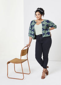Woman wearing a patterned cardigan and jeans stands confidently next to a wooden chair against a plain backdrop, conveying a casual and poised mood.