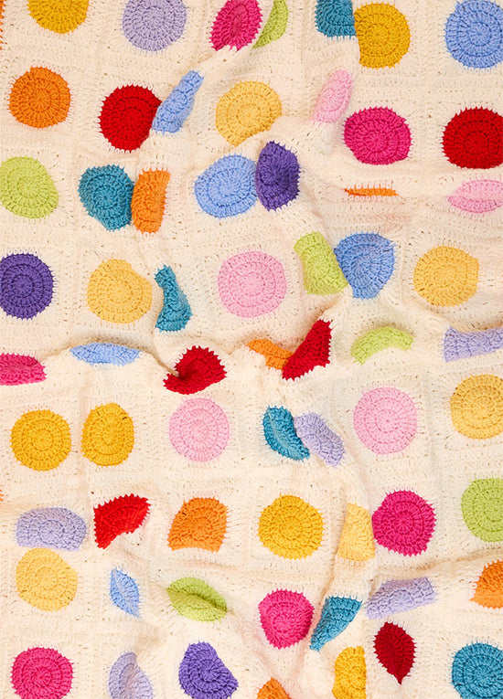 Crocheted Dot Blanket with colorful circular dots in red, yellow, blue, green, purple, pink, and orange on an off-white background – playful handmade design with a soft, slightly folded texture.