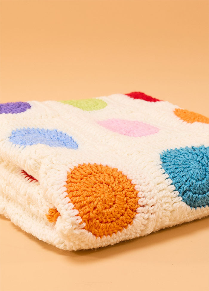 Folded crocheted Dot Blanket with a white base and colorful circular patterns in orange, blue, purple, green, pink, and red, displayed on a beige background – cheerful and handmade design.