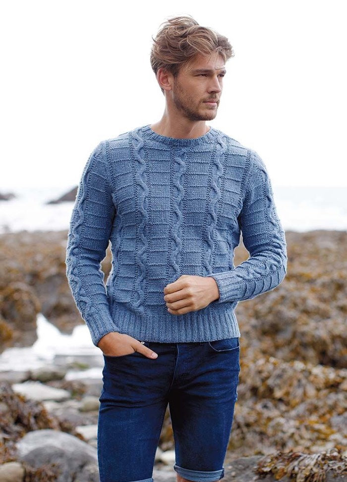 Young man in a blue cable-knit sweater and jeans stands on a rocky beach. He looks into the distance with a thoughtful expression, conveying a serene mood.