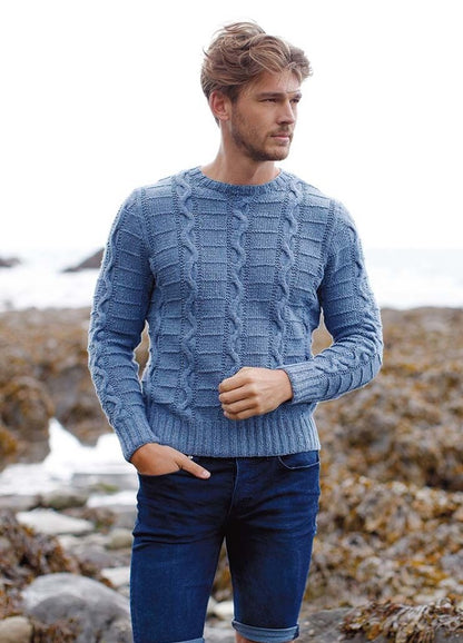Young man in a blue cable-knit sweater and jeans stands on a rocky beach. He looks into the distance with a thoughtful expression, conveying a serene mood.