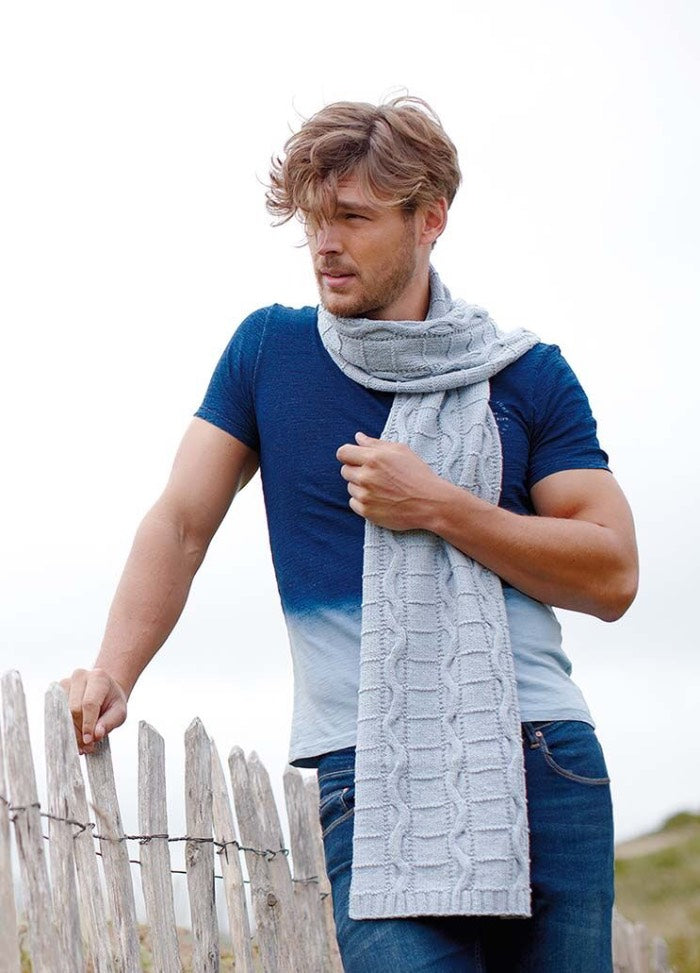 Man in a blue shirt and knitted gray scarf stands by a rustic wooden fence, facing a windy, cloudy sky. His pose is relaxed yet thoughtful.