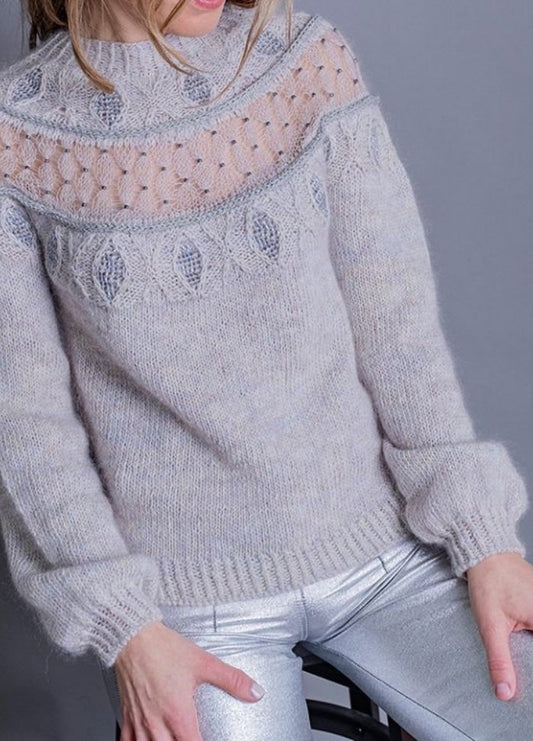 A person wearing a light gray knitted sweater with intricate patterns and sheer details at the top sits against a gray background, paired with shiny silver pants.